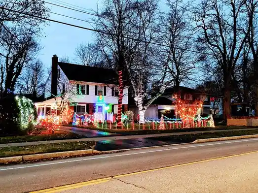 A residential house at dusk is covered in elaborate, colorful christmas lights, including trees, bushes, and decorative…