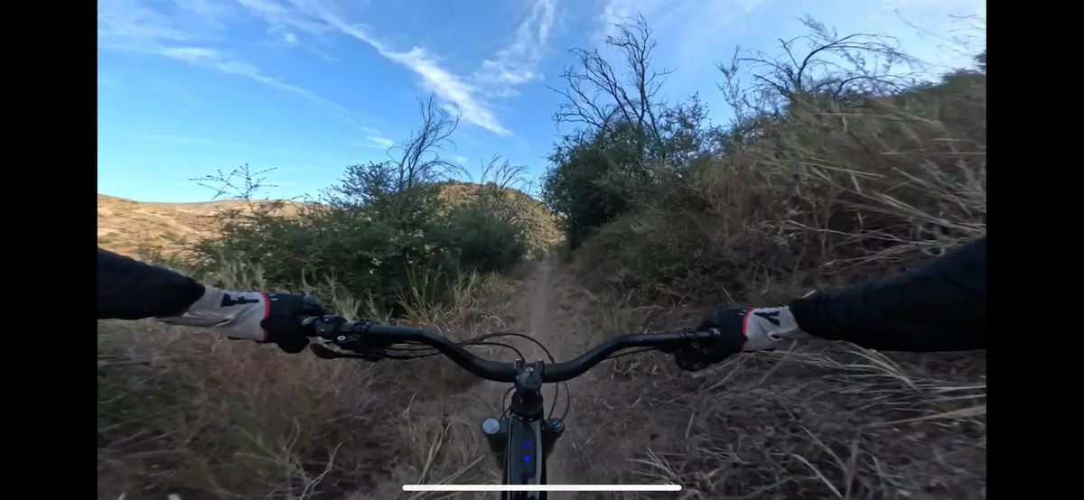 A first-person view from a mountain bike rider on a narrow dirt trail through dry brush and sparse trees under a bright blue…