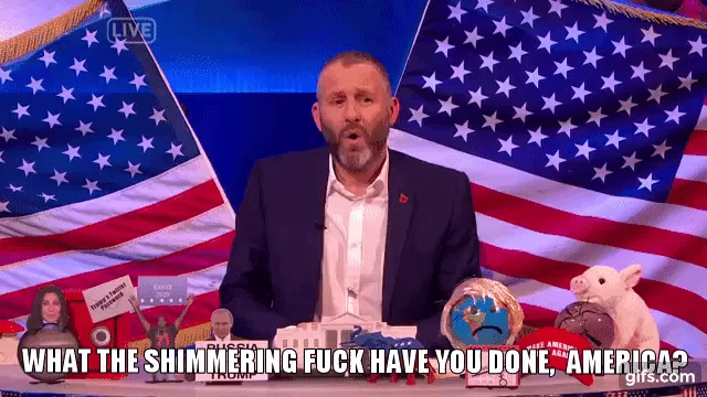 A man with a beard and dark suit sits at a desk in front of an American flag, visibly reacting with escalating shock and…
