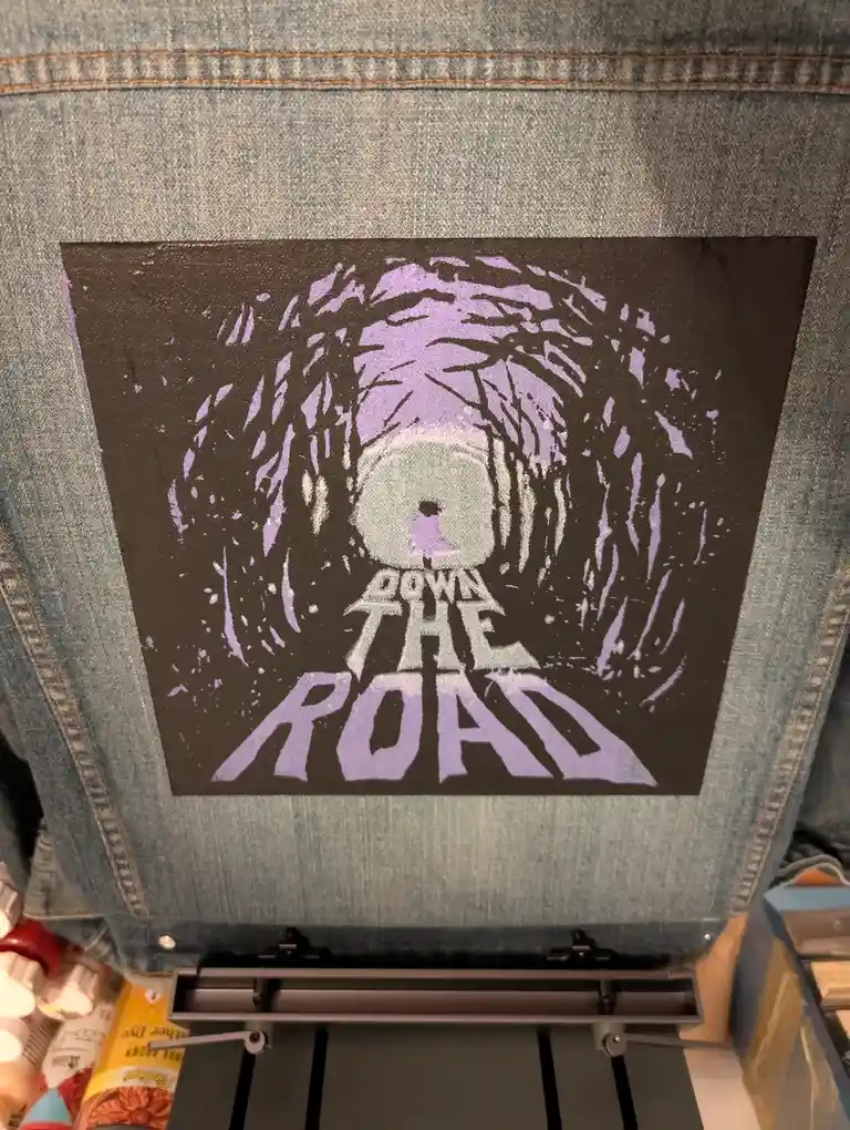 A denim jacket back features a stenciled graphic of a road stretching into a tunnel of trees, with the text 'DOWN THE ROAD'…