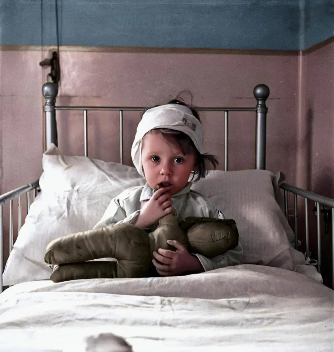 A young child with a bandaged head sits in a metal-framed hospital bed, clutching a worn stuffed doll while looking directly…
