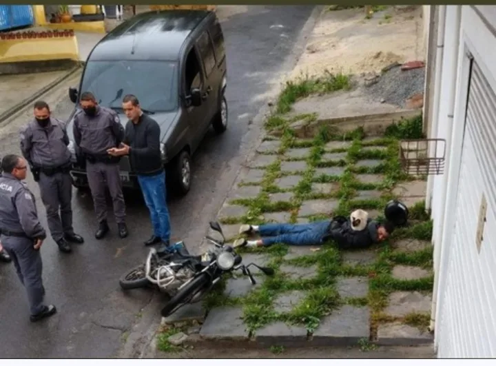 A man lies unconscious on a grassy sidewalk next to a fallen motorcycle, while several police officers and a civilian stand…