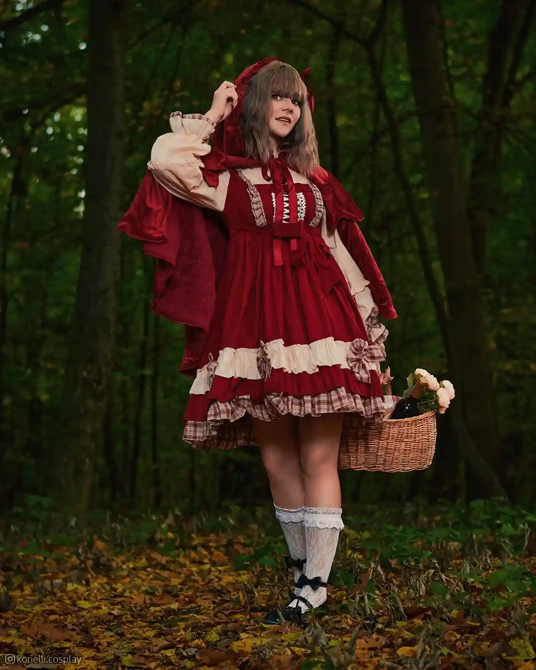 A person in a red hooded dress with ruffles and plaid accents stands in a forest holding a basket containing a bottle and…