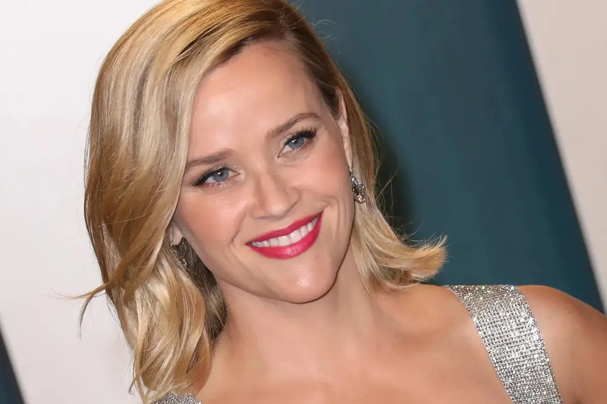 a close-up photo of actress Reese Witherspoon smiling warmly at the camera, wearing a sparkling silver dress and bright red…