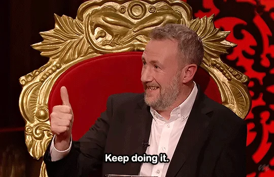 A man with a beard wearing a black suit and white shirt sits in a red and gold ornate chair, smiling and giving a thumbs-up…