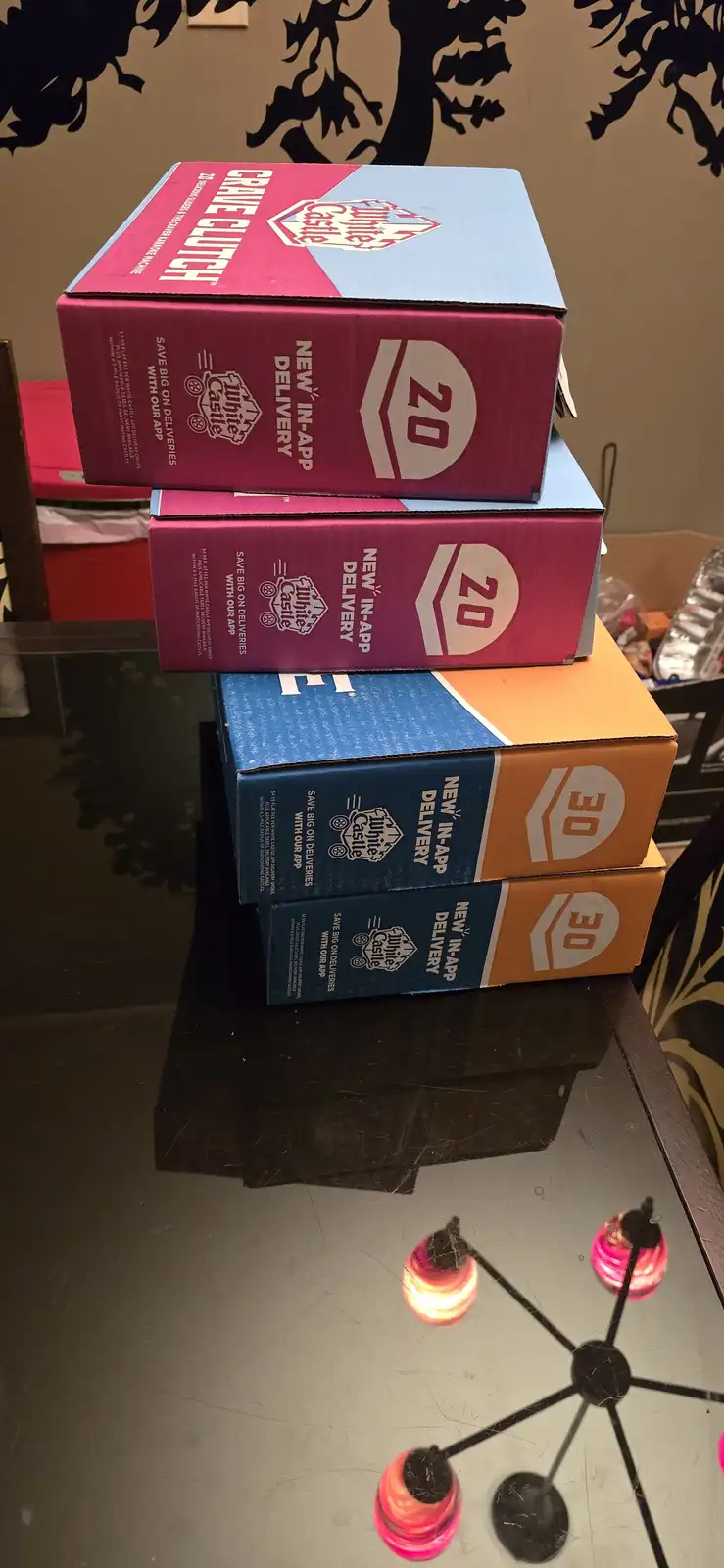 Four White Castle burger boxes are stacked on a glass table; two pink boxes labeled '20' and two blue/orange boxes labeled…