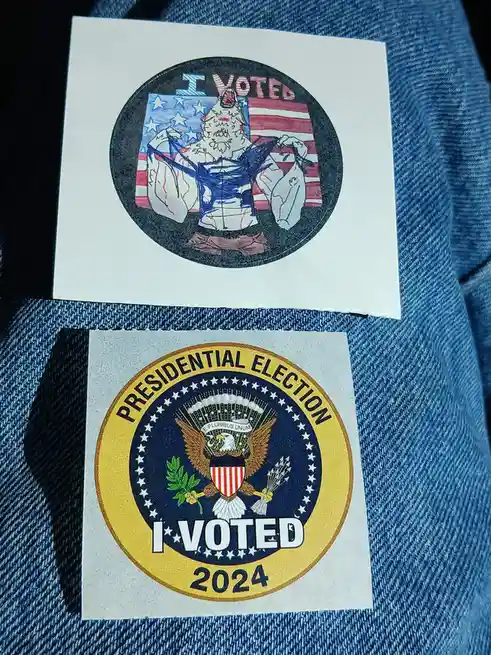 Two 'I VOTED' stickers are shown on denim fabric. The top sticker is a hand-drawn parody featuring a muscular, bloodied…