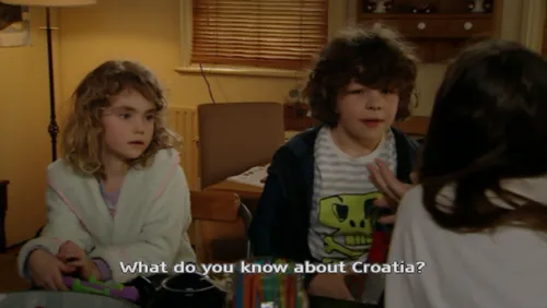 Three children are in a living room; a boy in a skull t-shirt is being asked a question while a girl in a robe looks on. The…