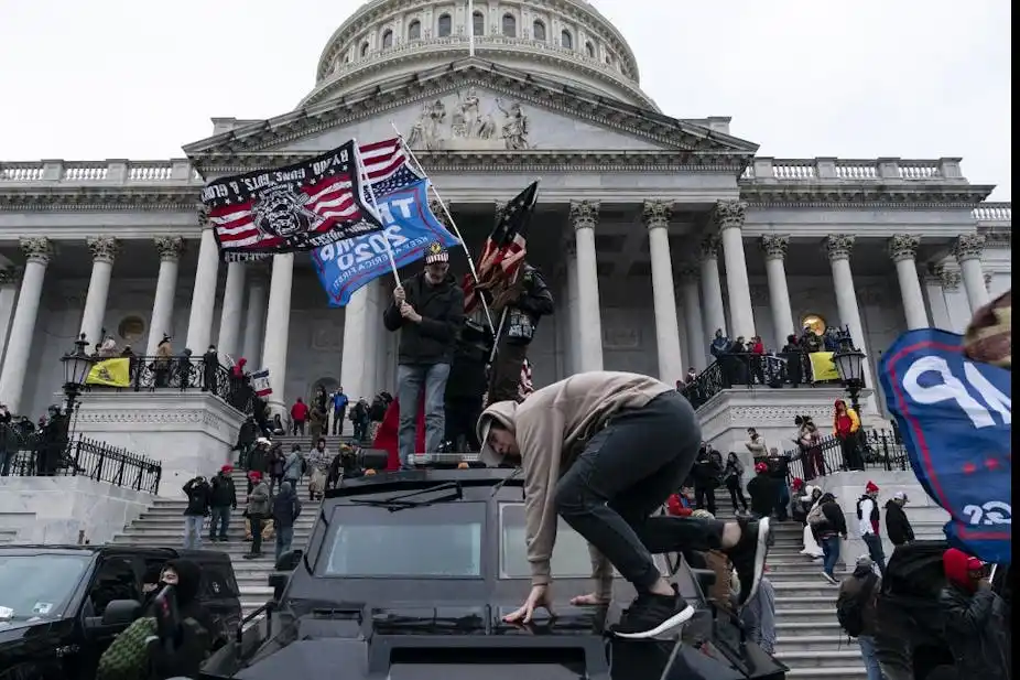 A person in a beige hoodie climbs onto the roof of a black armored vehicle in front of the U.S. Capitol building, while…