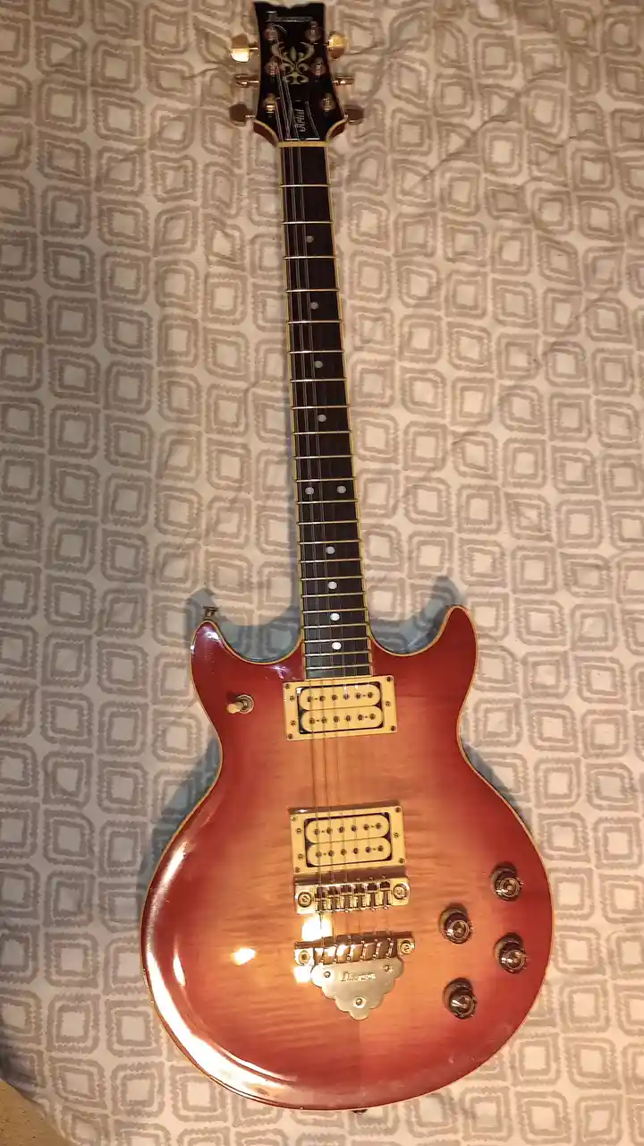 A red sunburst electric guitar with gold hardware lies flat on a patterned beige and gray fabric surface. The overlaid text…
