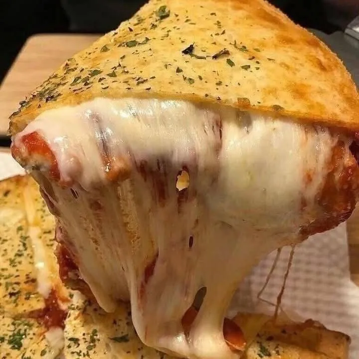 a close-up photo of a cheesy, breaded pizza slice with molten cheese stretching dramatically from the center, topped with…