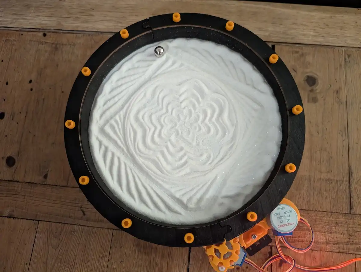 A top-down view of a circular black-framed device on a wooden surface, filled with white sand that has been arranged into a…