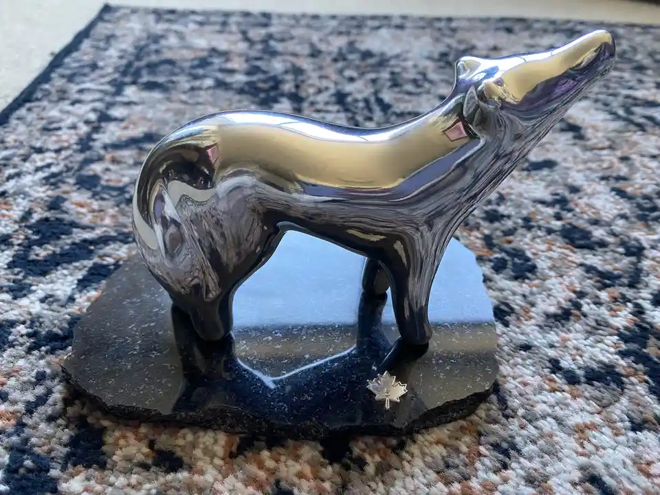 A shiny, chrome-finished wolf statue stands on a dark, rough-edged stone base, placed on a patterned rug. The statue's…