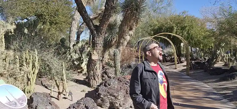 A man with glasses and a beard stands in a desert botanical garden, mouth wide open as if yelling, surrounded by various…