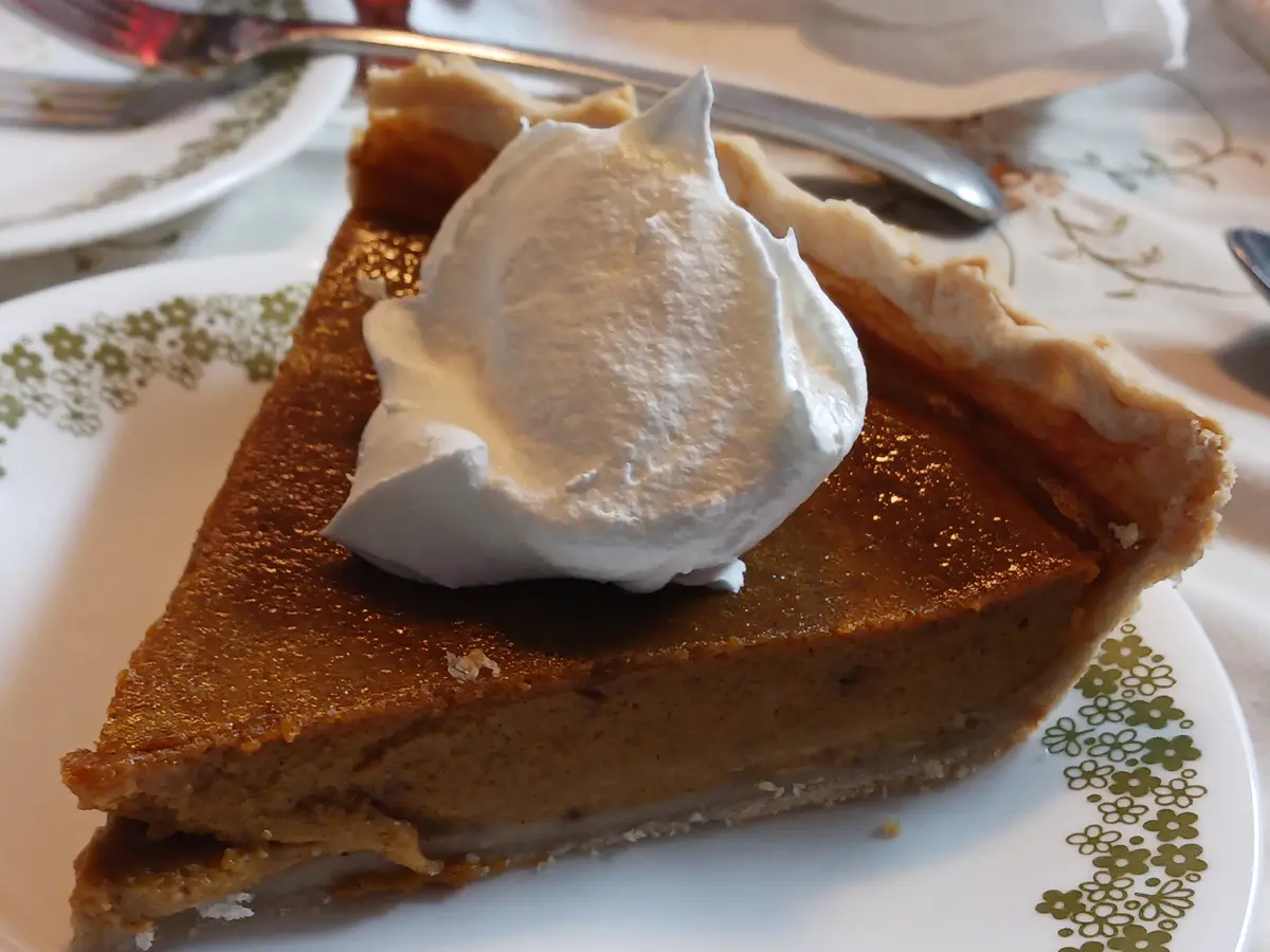 A close-up photo shows a slice of pumpkin pie with a dollop of whipped cream on top, served on a white plate with a green…