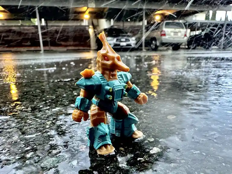 A small, orange and teal plastic action figure with a dinosaur-like head stands in a puddle under an overpass during a heavy…