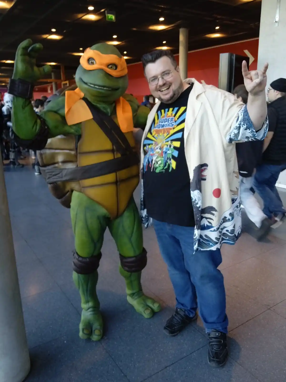 A man in a convention setting poses with a person in a full Teenage Mutant Ninja Turtles costume, specifically Michelangelo…