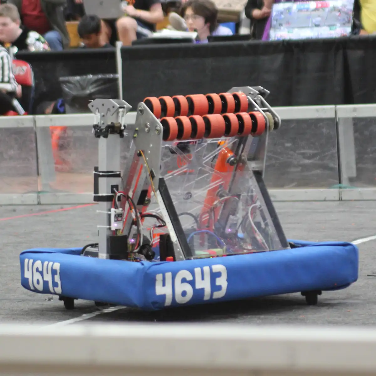A blue robot with the number 4643 on its sides is shown at a robot combat event, featuring a transparent body revealing…