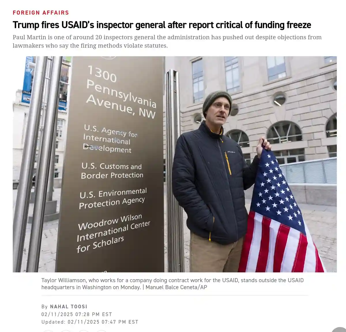 A man in a beanie and jacket stands outside a government building holding an American flag, next to a sign listing USAID and…