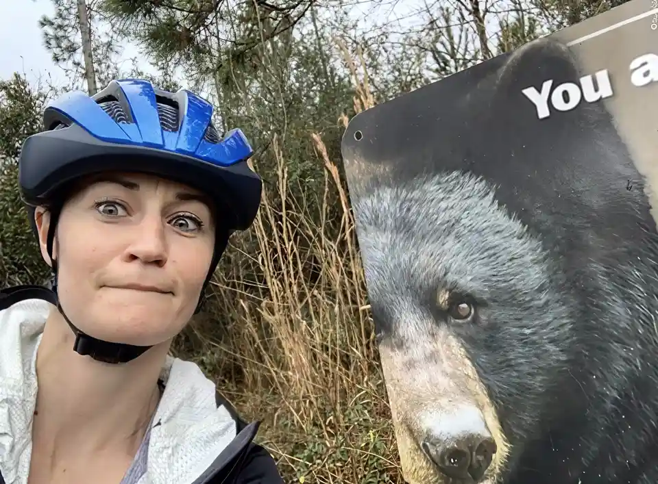 A person wearing a blue and black bicycle helmet takes a selfie outdoors next to a large sign featuring a close-up photo of…
