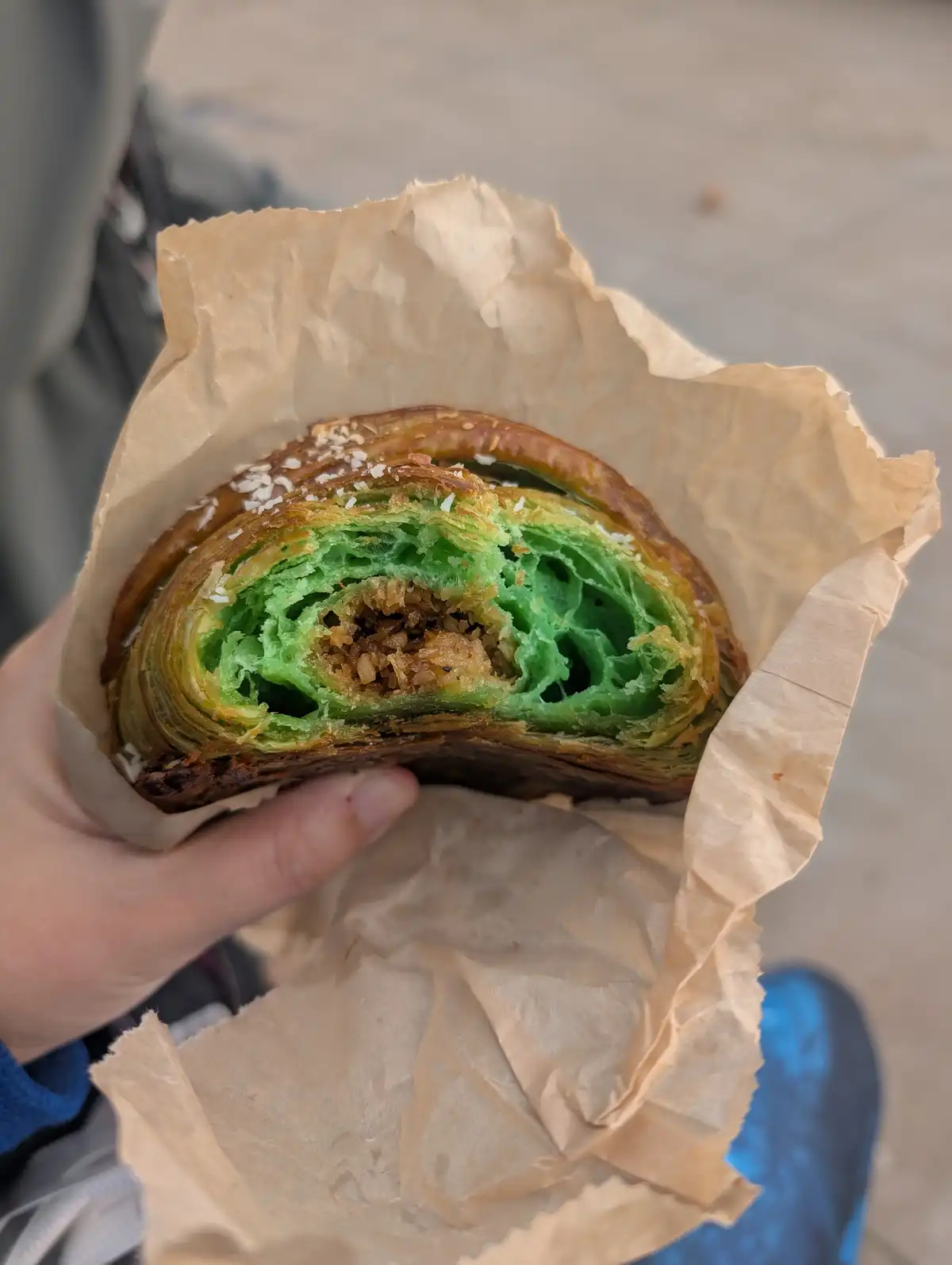 A hand holds a partially eaten, flaky pastry wrapped in brown paper, revealing a bright green interior and a brown filling…