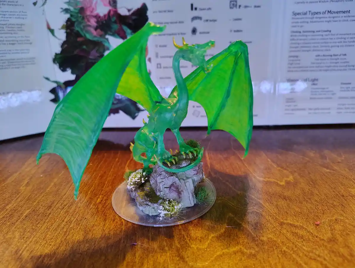 A translucent green dragon miniature with spread wings stands on a rocky, grassy base atop a wooden table. Behind it, an…