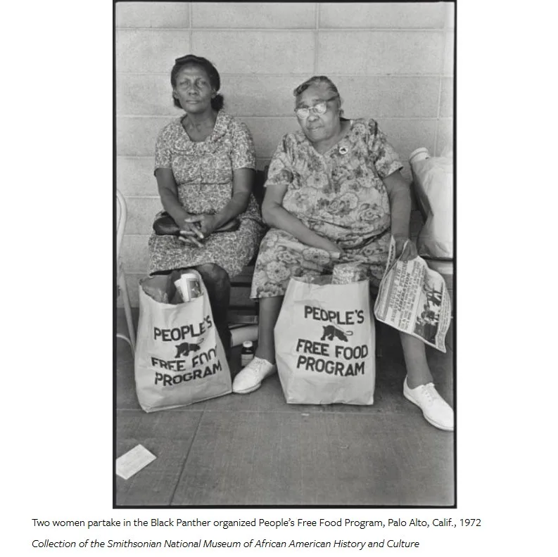 Two Black women sit on a bench next to large paper bags labeled 'PEOPLE'S FREE FOOD PROGRAM' from the Black Panther Party…