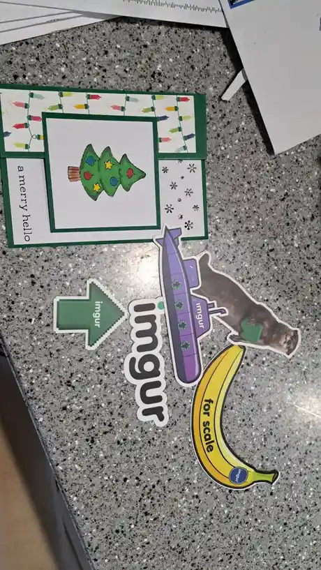 A handmade Christmas card with a decorated tree and "a merry hello" text is shown next to a banana, an otter holding a green…