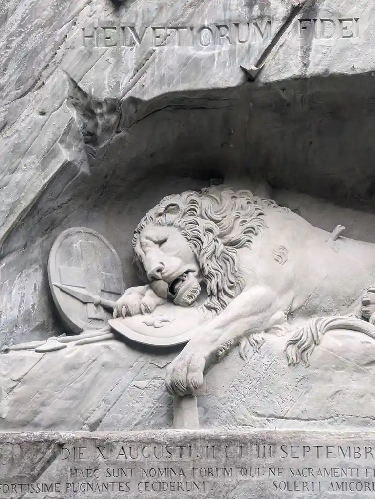 A stone relief depicts a carved lion lying down with its mouth open, holding a shield in its paw, next to a shield and…