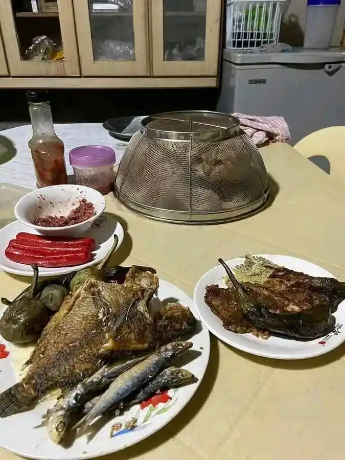 A kitchen table is set with plates of cooked fish, small fish, eggplant, and red peppers, alongside condiments. A metal mesh…