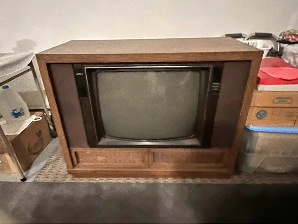 A vintage wooden-cased CRT television sits in a cluttered storage area surrounded by boxes and household items. The screen…