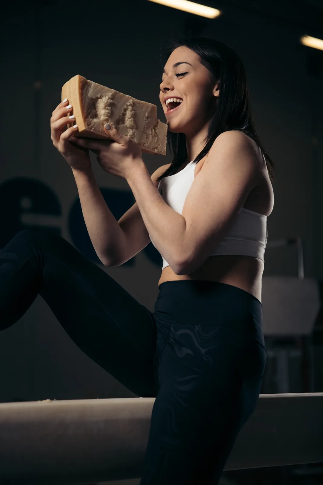 A woman in athletic wear, sitting on a balance beam in a gym, holds a large wedge of hard cheese with a joyful, exaggerated…