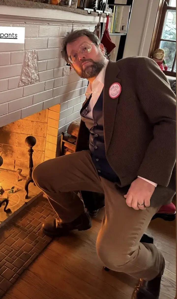 A man with glasses and a beard, wearing a brown jacket, vest, and pants, is kneeling beside a lit fireplace in a cozy room…
