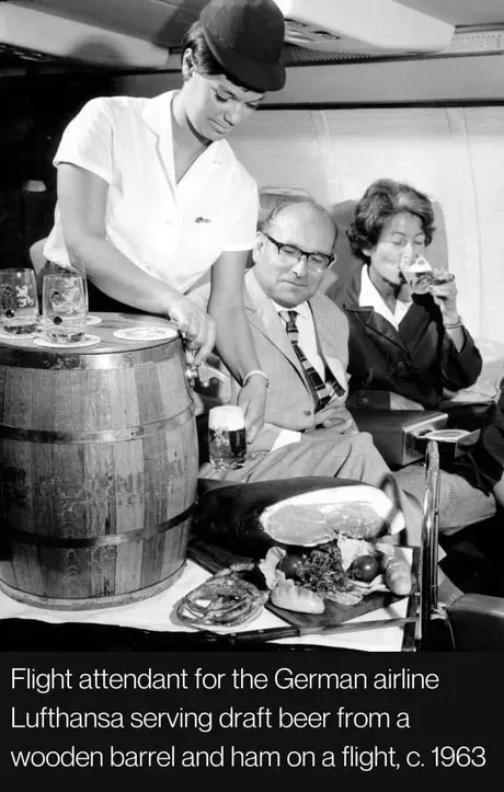 A black-and-white photo from circa 1963 shows a Lufthansa flight attendant pouring draft beer from a wooden barrel onto a…