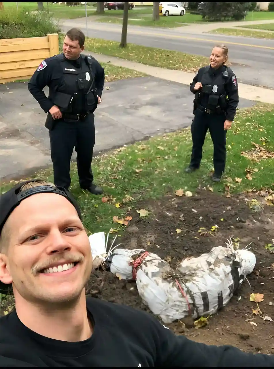 A man takes a selfie smiling broadly, with two police officers standing behind him in a residential yard. In the foreground…
