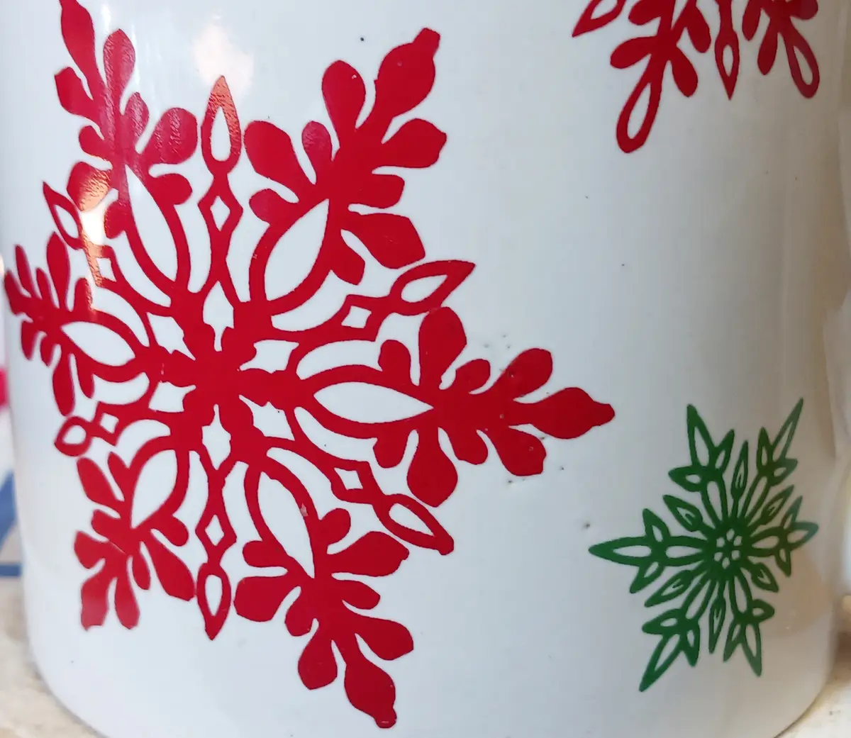 a close-up of a white ceramic mug with red and green snowflake designs; the red snowflake is large and ornate, while a…