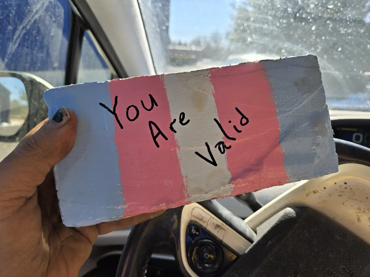 A hand holds a handmade sign painted with the pink, white, and blue stripes of the transgender flag, with the words 'You Are…
