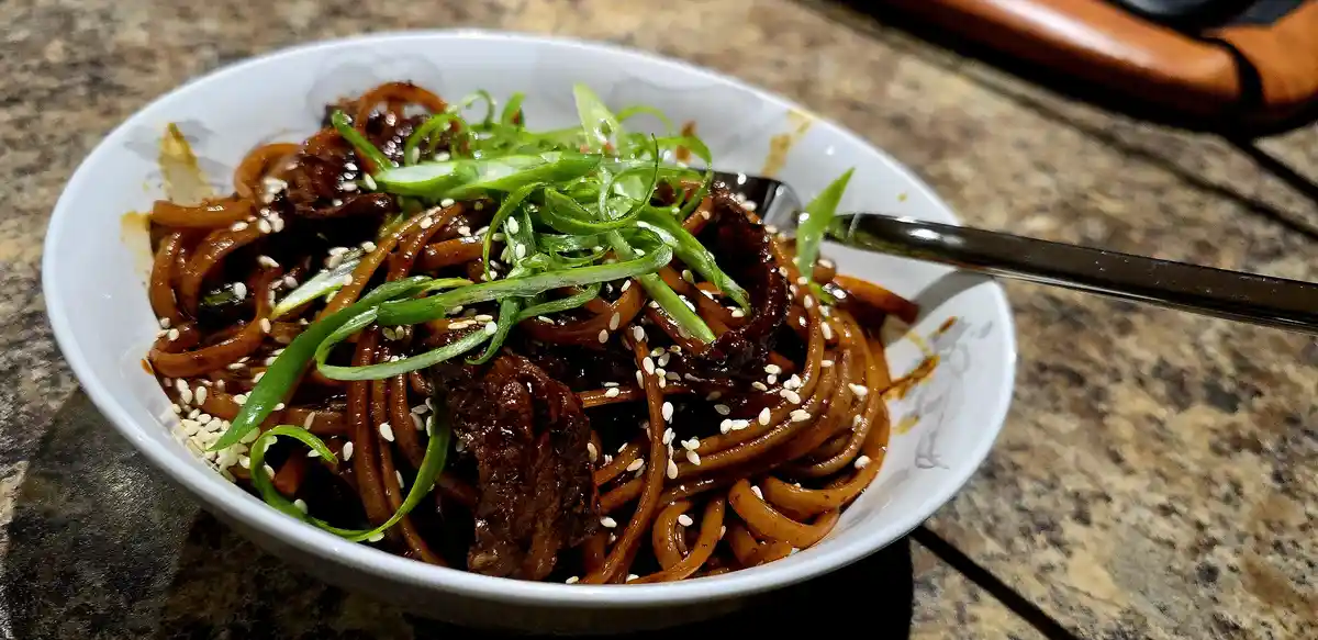A close-up photo of a bowl of glossy, dark sauce-coated noodles with sliced meat, topped with chopped green onions and white…