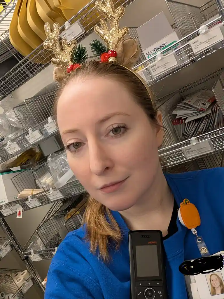 A woman in blue scrubs wearing a glittery reindeer antler headband smiles at the camera while holding an ascom phone in a…