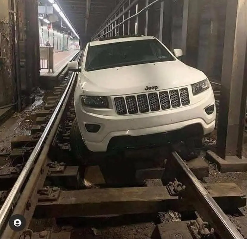 A white Jeep Grand Cherokee is parked directly on a set of subway tracks inside a station, with its front wheels resting on…