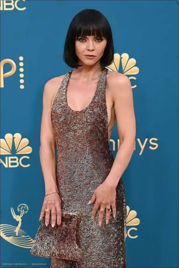 A woman with a short black bob haircut and bangs stands at a red carpet event, wearing a shimmering, textured halter-neck…