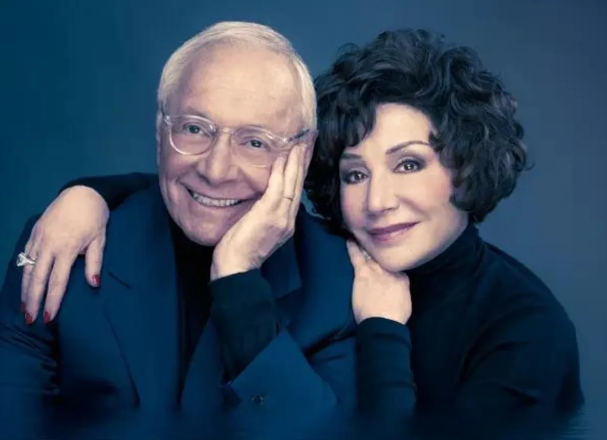 An older man and woman are posing together in a studio portrait against a blue background. The woman has her arm around the…