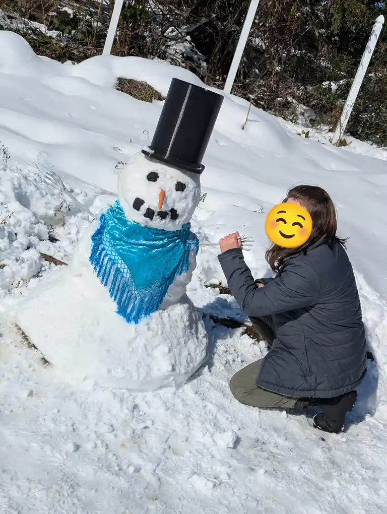 A person with a smiling emoji covering their face kneels in snow next to a snowman wearing a black top hat and a shiny blue…