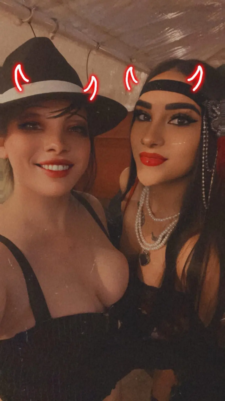 Two women pose for a selfie in a dimly lit setting, both with glowing red devil horns digitally added above their heads. The…