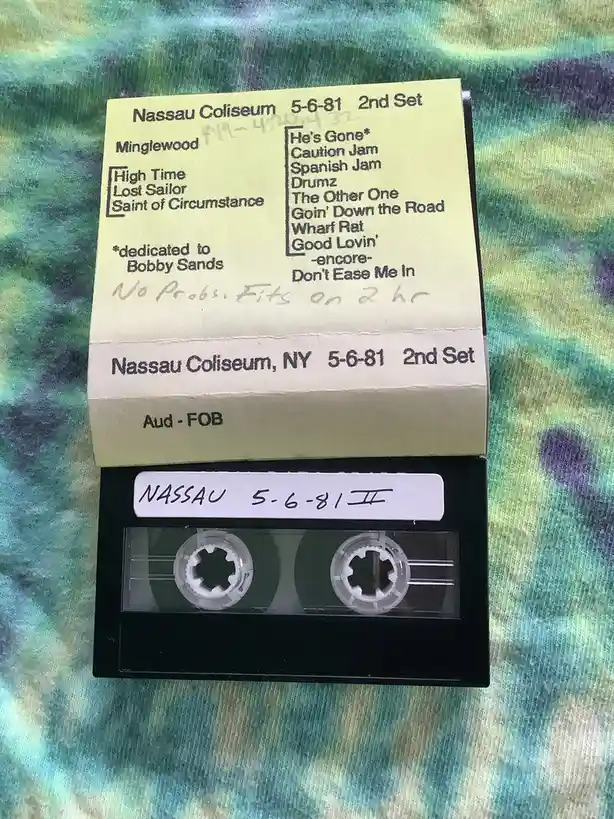 A black audio cassette tape with handwritten and printed labels sits on a tie-dye fabric. The top label lists a Grateful…