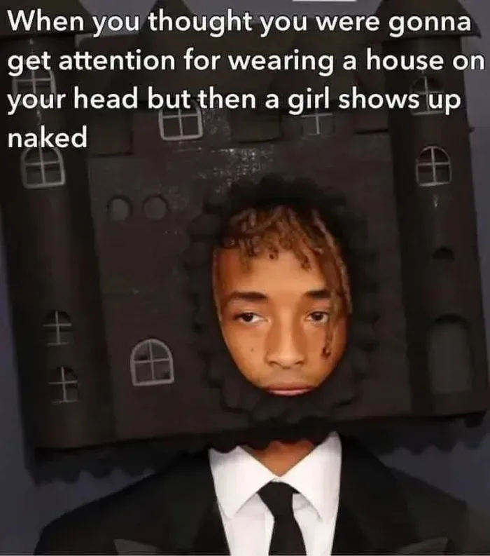 A person wearing a black cardboard house on their head, with windows and turrets, while dressed in a formal black suit…