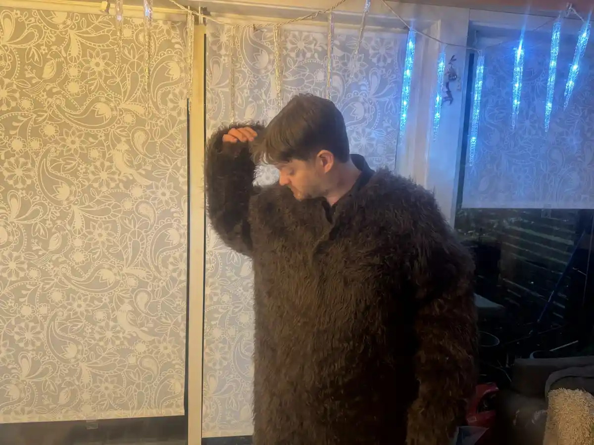 A man in a dark, shaggy, fur-like coat stands indoors, looking down and touching his hair with one hand. Behind him are…