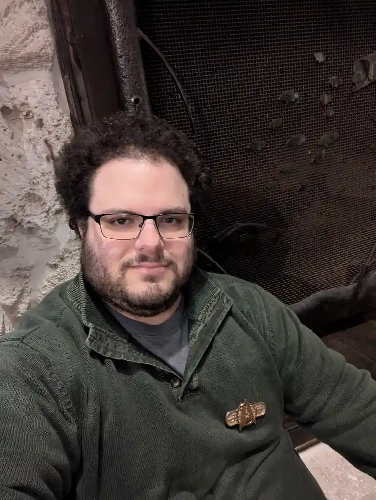 A man with curly dark hair, glasses, and a beard wears a green corduroy jacket over a gray shirt. A gold Starfleet insignia…