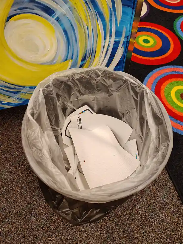 A trash bin lined with a plastic bag contains crumpled graph paper with faint drawings, positioned next to a colorful…