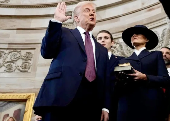A man in a dark suit and purple tie raises his right hand while speaking, standing beside a woman in a dark hat holding a…